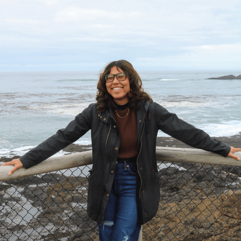Young woman standing in front of an ocean view. She is wearing glasses, a long black jacket, a brown sweater, and dark blue jeans.